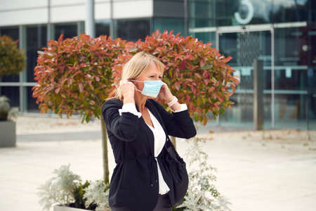 Mature Businesswoman Putting On PPE Face Mask Walking Outdoors In Street During Health Pandemicの写真素材