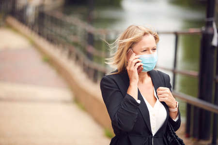 Mature Businesswoman With Mobile Phone Wearing PPE Face Mask Walks In Street During Health Pandemicの写真素材