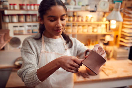 Female Potter Fitting Clay Handles To Mugs In Ceramics Studio