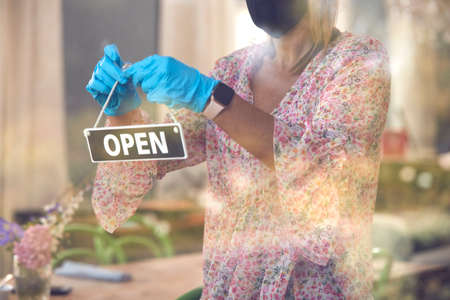 Female Owner Of Coffee Shop Wearing Face Mask Turning Round Open Sign During Health Pandemic