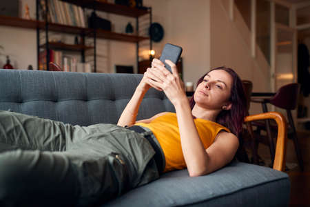 Young Woman Lying On Sofa At Home Looking At Mobile Phone Messages And Social Mediaの写真素材