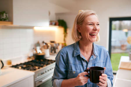 Laughing Mature Woman At Home In Kitchen Drinking Coffeeの写真素材