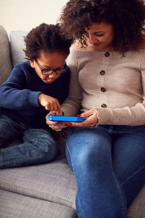 Mother And Son Sitting On Sofa At Home Playing Computer Game Together On Hand Held Device At Home