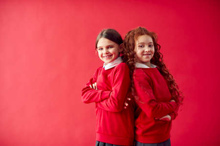 Two Elementary School Pupils Wearing Uniform Back To Back Against Red Studio Background
