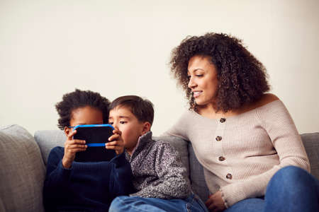Mother And Sons Sitting On Sofa At Home Playing Computer Game Together On Hand Held Device At Home