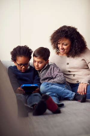 Mother And Sons Sitting On Sofa At Home Playing Computer Game Together On Hand Held Device At Home
