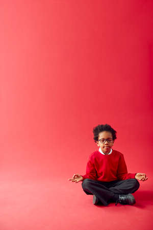 Male Elementary School Pupil Wearing Uniform Sitting And Meditating Against Red Studio Backgroundの写真素材