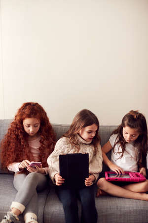 Group Of Girls With Friends Sitting On Sofa At Home Playing On Digital Tablet And Mobile Phonesの写真素材
