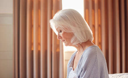 Senior Woman With Serious Expression Sitting On Edge Of Bed At Home During Lockdown For Covid-19
