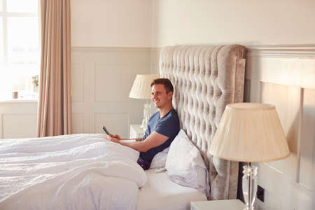 Man At Home In Bed Self Isolating Using Digital Tablet And Mobile Phone During Covid 19 Lockdown