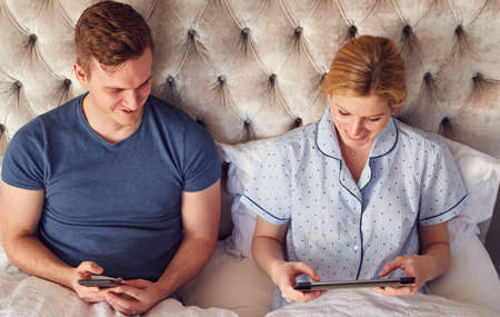 Couple At Home In Bed Self Isolating Using Digital Tablet And Mobile Phone During Covid 19 Lockdown
