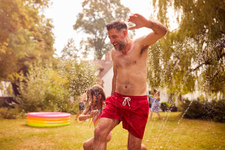 Father And Daughter Run Through Water From Garden Sprinkler Having Fun Wearing Swimming Costumes