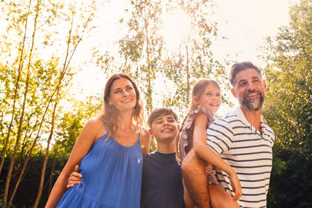 Portrait Of Smiling Family In Summer Garden With Father Giving Daughter Piggyback Ride On Back
