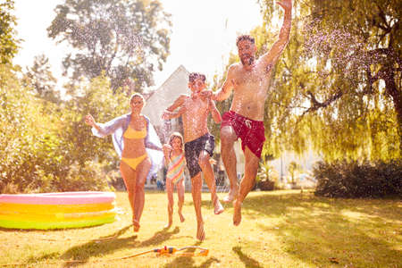 Family Running Through Water From Garden Sprinkler Having Fun Wearing Swimming Costumesの写真素材