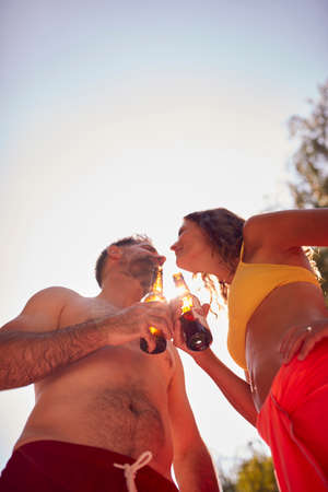 Couple Wearing Swimming Costumes Standing In Paddling Pool In Summer Garden At Home Drinking Beer