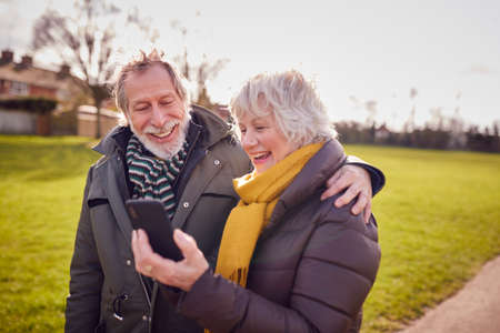 Loving Senior Couple Looking At Mobile Phone Enjoying Autumn Or Winter Walk Through Park Togetherの写真素材