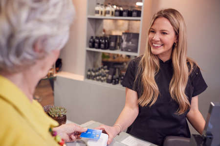 Senior Woman Making Contactless Payment To Stylist In Salon With Credit Card