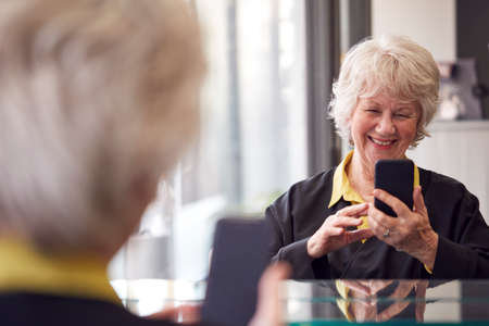 Senior Woman Waiting To Have Hair Cut In Hairdressing Salon Looking At Mobile Phone