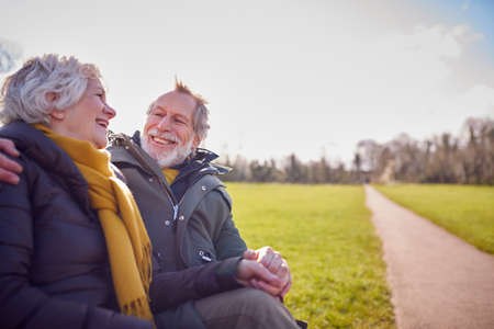 Loving Senior Couple Sitting On Seat Enjoying Autumn Or Winter Walk Through Park Togetherの写真素材