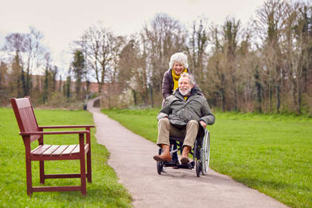 Senior Woman Pushing Senior Man In Wheelchair Outdoors In Fall Or Winter Parkの写真素材