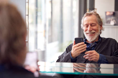 Senior Man Waiting To Have Hair Cut In Hairdressing Salon Looking At Mobile Phone