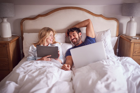 Couple Lying In Bed In Morning Looking At Digital Tablet And Laptop