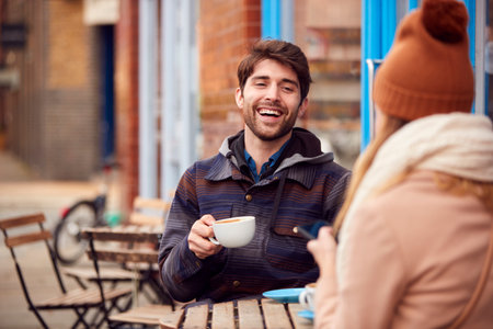 Couple On Date Sitting Outside Coffee Shop On Busy City High Street Looking At Digital Devicesの写真素材