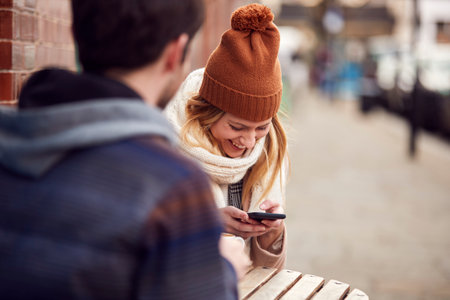 Couple On Date Sitting Outside Coffee Shop On High Street Using Mobile Phoneの写真素材