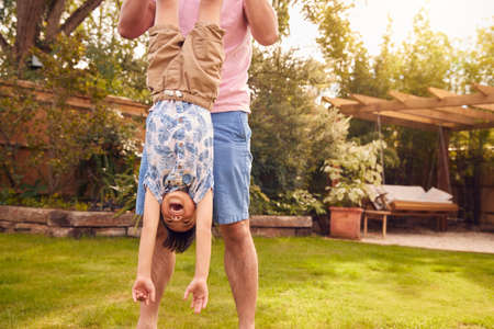 Asian Father Holding Son Upside Down By Legs In Garden As They Play Game Together