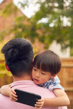 Asian Father Cuddling Son In Garden As Boy Looks Over His Shoulder At Mobile Phoneの写真素材