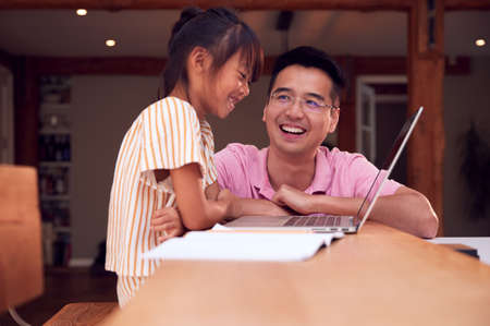 Asian Father Helping Home Schooling Daughter Working At Table In Kitchen On Laptopの写真素材