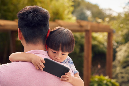 Asian Father Cuddling Son In Garden As Boy Looks Over His Shoulder At Mobile Phoneの写真素材