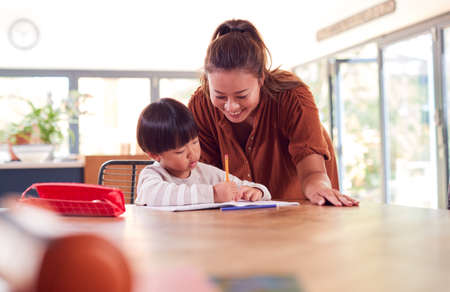 Asian Mother Helping Home Schooling Son Working At Table In Kitchen Writing In Bookの写真素材