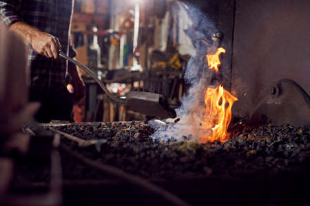 Male Blacksmith Adding Coal Fuel To Wood Kindling To Start Blaze In Forge