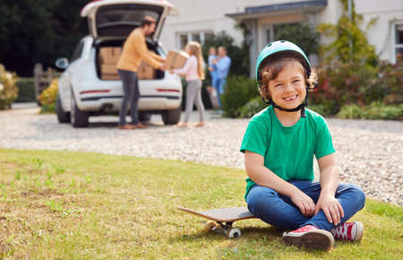 Portrait Of Boy With Skateboard Outside New Family Home On Moving Day Unloading Boxes From Car