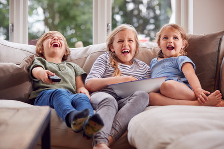 Three Children Sitting On Sofa At Home Laughing And Watching TV With Popcornの写真素材