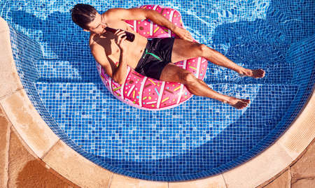 Overhead Shot Of Man In Swim Shorts Floating In Ring Drinking Beer In Outdoor Swimming Poolの写真素材