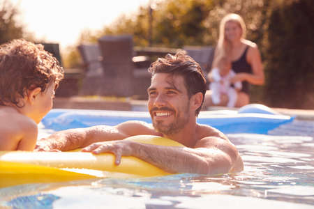 Father And Son Have Fun Playing In Outdoor Pool On Vacation As Mother And Baby Watch From Sideの写真素材
