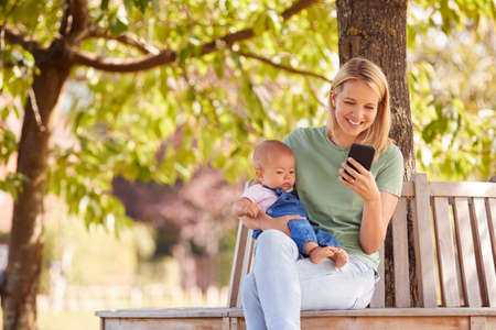 Mother Sitting On Park Bench Under Tree With Baby Daughter Looking At Mobile Phone