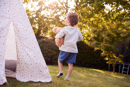 Young Boy Carrying Teddy Bear Having Fun With Tent Or Tepee Pitched In Gardenの写真素材