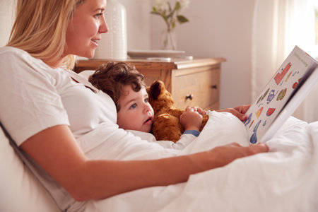 Mother In Bedroom Looking At Picture Book With Young Son Wearing Pyjamas Cuddling Soft Toyの写真素材