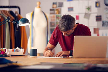 Mature Male Fashion Designer In Studio Working On Sketches Or Documents At Desk With Laptopの写真素材