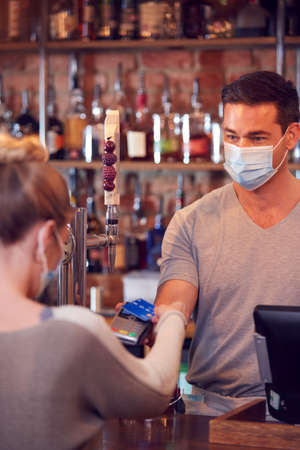 Female Customer Wearing Mask In Bar Making Contactless Payment For Drinks During Health Pandemicの写真素材