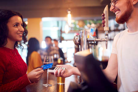 Smiling Female Customer In Bar Making Contactless Payment With Card For Drinks To Male Bartenderの写真素材