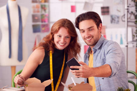 Male And Female Fashion Business Owners Having Working Lunch Looking At Mobile Phone In Studioの写真素材