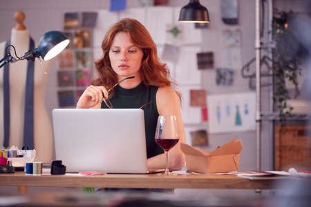 Female Owner Of Fashion Business With Glass of Wine Working Late On Laptop In Studio