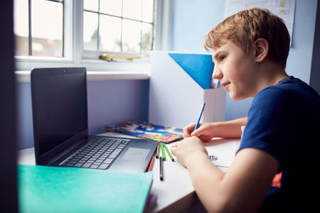 Boy Sitting At Desk Home-Schooling Using Laptop For Online Learning During Health Pandemicの写真素材