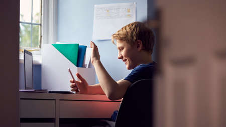 Boy Sitting At Desk Home-Schooling Making Online Video Call On Mobile Phone And Using Laptopの写真素材