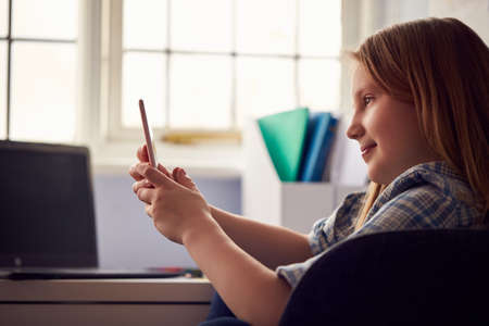 Girl Sitting At Desk Home-Schooling Making Online Video Call On Mobile Phone And Using Laptopの写真素材