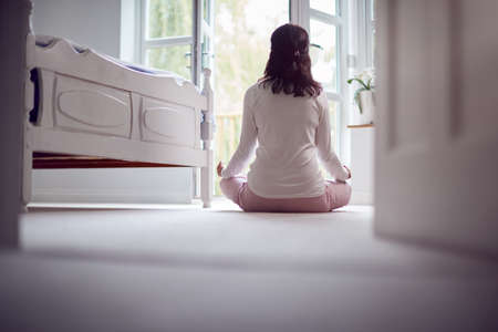 Rear View Of Mature Asian Woman In Pyjamas Sitting On Bedroom Floor Meditating In Yoga Poseの写真素材
